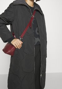 Abrigo largo acolchado negro con cierre frontal y gran cuello. Bolso cruzado rojo con forma estructurada y correa ajustable.