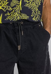 Pantalones cortos de mezclilla negra con cintura elástica, detalle de cordón, bolsillos delanteros y acentos desgarbados. Combinados con una camisa de patrón multicolor.