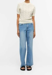 Suéter de punto crema de manga corta con un patrón texturizado de diamantes, combinado con pantalones vaqueros de pierna ancha azul claro y sandalias negras.