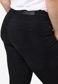 Primer plano de una persona que lleva jeans negros y una camiseta blanca, mostrando el bolsillo trasero y la zona de la cinturilla.