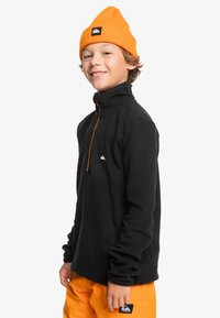 Schwarzer Fleece-Pullover mit hohem Kragen und einem orangefarbenen Reißverschluss. Kombiniert mit leuchtend orangen Hosen. Mit einem Logo auf der Brust und dem Saum.