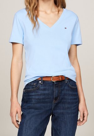 Persoon draagt een lichtblauw T-shirt met V-hals met een klein logo, donkerblauwe jeans en een bruine riem, staand tegen een neutrale achtergrond.