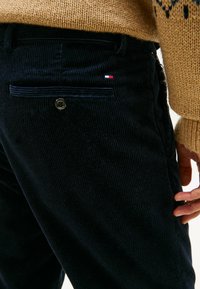 Navy corduroy-housut, joissa on ribbed-rakenne. Sahan takana nappikiinnitys ja pieni brodeerattu logo punaisella, valkoisella ja sinisellä.
