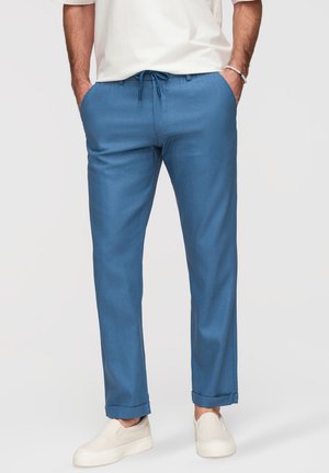 Hombre con pantalones azules con cordón, remangados en los puños, con zapatos blancos tipo slip-on y manos en los bolsillos, sobre un fondo liso.