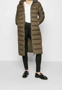 Olivgrön, lång pufferjacka med horisontella quiltade sektioner, huva och dragkedjestängning. Kombinerad med svarta tajta byxor och loafers.