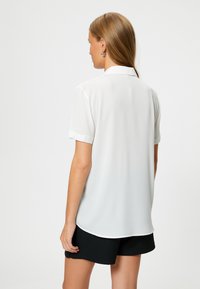Blusa bianca a maniche corte con colletto, texture liscia e orlo arcuato. Indossata con pantaloni corti neri, vista da dietro.