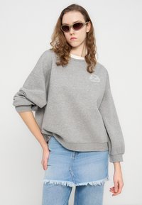 Γκρι φούτερ με ριμπ μανσέτες, διακοσμημένο με ένα μίνιμαλ κεντητό σχέδιο. Συνδυάζεται με μίνι φούστα από τζιν με φθαρμένο τελείωμα.