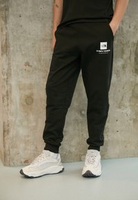 Svarta bomullsträningbyxor med avsmalnande ben och ankle cuffs, med en vit logopatch och geografiska koordinater på fickan.