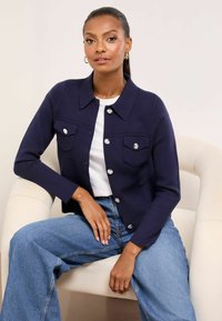 Tengerészkék kötött dzseki gallérral, ezüst gombos záródással és két mellzsebbel, fehér inggel és kék, széles szárú farmerrel párosítva.