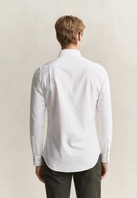 Homme aux cheveux blonds courts, portant une chemise blanche à manches longues et un pantalon sombre, debout de dos sur un fond uni.