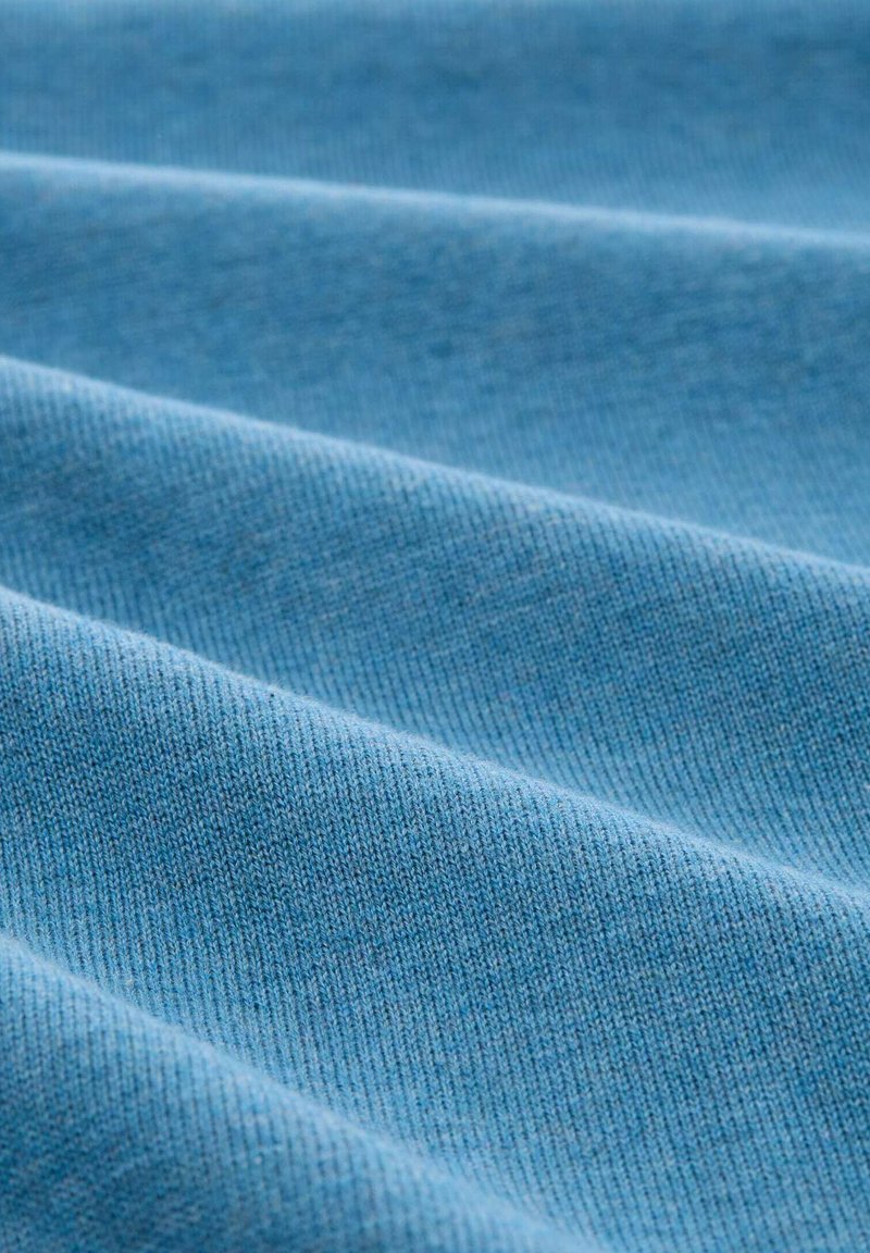 Tessuto azzurro chiaro con una superficie testurizzata, che presenta sottili linee diagonali e pieghe, suggerendo un materiale morbido e traspirante.