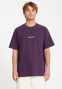 Lilla bomulds t-shirt med korte ærmer, rund hals, prydet med et hvidt "VOLCOM" logo på brystet. Modellen bærer lyse bukser.