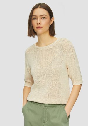 Pull en tricot à manches courtes de couleur beige clair, avec un col rond et un tissu texturé. Porté avec un pantalon vert olive, les mains dans les poches.