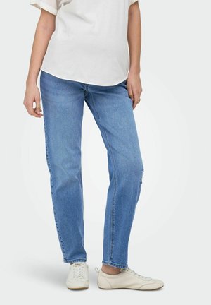 Nő világoskék egyenes szárú farmerral, fehér fűzős cipővel és törtfehér rövid ujjú felsővel, egyszínű háttér előtt áll.