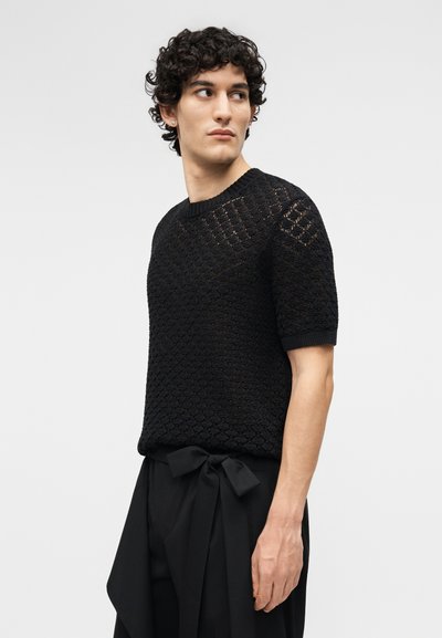 Νεαρός με σγουρά μαλλιά που φοράει μαύρο πλεκτό πουλόβερ με κοντά μανίκια και μαύρο παντελόνι με φιόγκο στη μέση, κοιτάζει προς τα πλάγια.