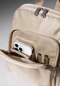Zaino in nylon beige con chiusura a doppia cerniera, dotato di una tasca esterna che contiene uno smartphone e due penne in supporti elastici.