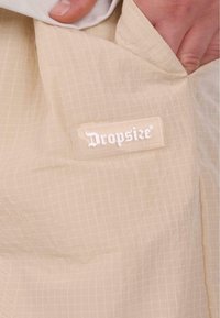 Tissu beige avec un motif en grille ; étiquette visible indiquant "Dropsize" en blanc. Matériau léger, légèrement texturé, avec des bords lisses.