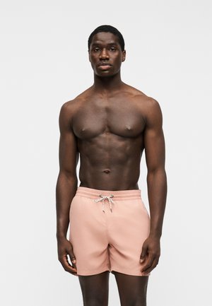 Jeune homme musclé à la peau foncée portant un short de bain rose clair avec cordon, debout devant un fond clair uni.