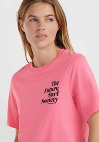 Růžové bavlněné tričko s černým grafikou, která obsahuje text "The Future Surf Society" a logo umístěné na přední straně.