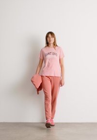 T-shirt in cotone rosa con testo "GANT 1949 NEW HAVEN", abbinato a pantaloni della tuta rosa coordinati e sneakers. Texture liscia e vestibilità rilassata.