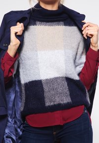 Person som bär en flerfärgad rutig stickad tröja över en röd skjorta och en mörkblå kappa, håller kappan öppen för att avslöja tröjan.