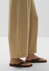 Beige linen trousers with a wide, flowing shape and visible seams at the hem, paired with dark brown flip-flop sandals featuring crisscross straps.