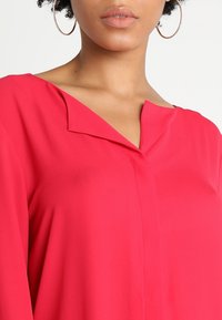 Blusa rosa brillante con scollatura a V e maniche lunghe. Realizzata in un tessuto morbido, presenta cuciture sottili sulla parte anteriore.