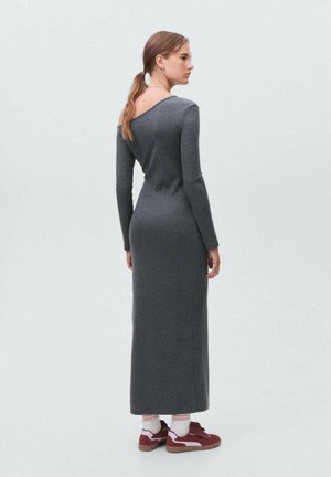 Graues, geripptes Langarmkleid mit einem schulterfreien Ausschnitt. Figurbetonte Silhouette mit einem geraden, knöchellangen Rock. Trägt rote Turnschuhe und weiße Socken.