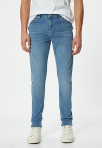 Vaqueros de corte Slim en un tono azul claro, fabricados en denim, con cinco bolsillos, una textura suave y un cierre de botón en la cintura. Combinados con zapatillas blancas.