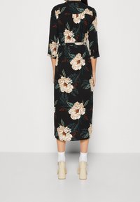 Czarna midi sukienka w kwiaty z beżowym i zielonym nadrukiem, z rękawami do łokci, talią ściągniętą i zapinana w pasie. Noszona z jasnymi butami.