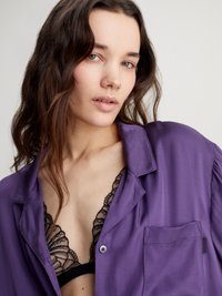 Cămașă de satin mov cu un buzunar pe piept, purtată peste un bralette din dantelă neagră. Materialul este neted, cu un luciu subtil și o textură moale.