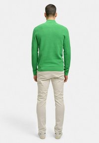 Grön texturerad stickad tröja med ribbad fåll, i kombination med ljusa byxor och neutrala sneakers; sedd bakifrån.