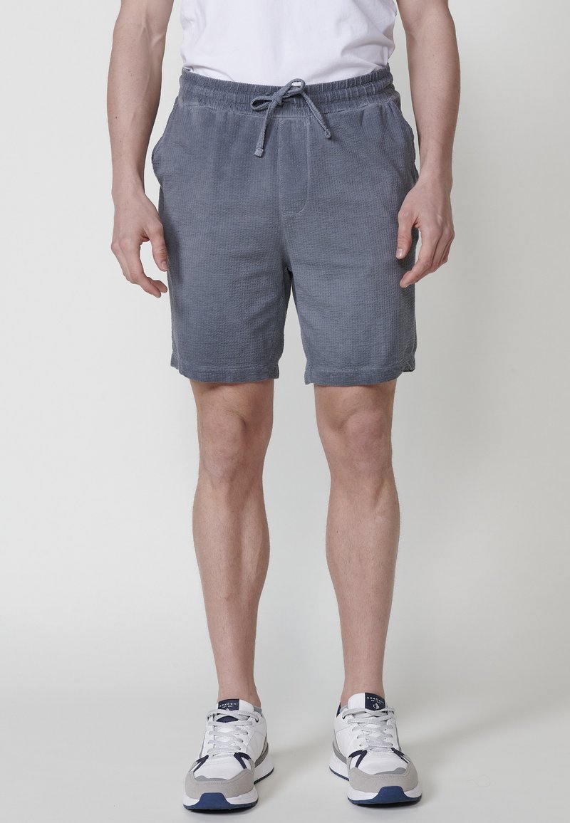 Uomo che indossa pantaloni corti grigi con coulisse, sneaker bianche e una maglietta bianca, in piedi contro uno sfondo chiaro e neutro.