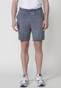 Man som bär gråa träningsoverallsbyxor, vita sneakers och en vit t-shirt, stående mot en enkel ljus bakgrund.