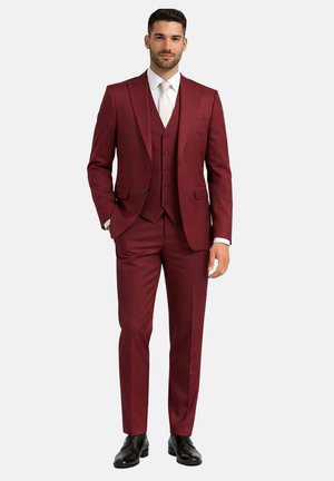 Homme portant un costume trois pièces bordeaux ajusté avec une chemise blanche, une cravate assortie et des chaussures de ville noires, debout avec une main dans la poche.