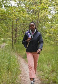 Giacca nera leggera sopra un felpa grigia, pantaloni rosa e scarpe da ginnastica. Ambientazione esterna con alberi e un sentiero di erba.