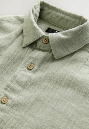 Chemise boutonnée texturée vert clair avec un col pointu et des boutons en bois, arborant un motif à carreaux subtil. Fini en tissu doux.