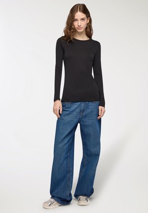 Donna con lunghi capelli mossi, indossa un top lungo aderente nero, jeans blu larghi e sneakers bianche, in piedi davanti a uno sfondo semplice.