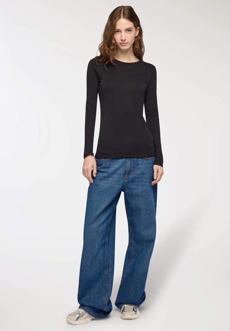 Donna con lunghi capelli mossi, indossa un top lungo aderente nero, jeans blu larghi e sneakers bianche, in piedi davanti a uno sfondo semplice.