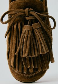 Brun ruskindsloafer med kvaster og en bundet sløjfe på den øverste del, der viser detaljeret tekstur og syninger.