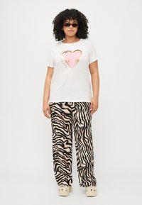 Un tricou alb cu un grafic în formă de inimă roz și litere aurii, purtat cu pantaloni bej cu un model de zebră neagră și saboți crem.