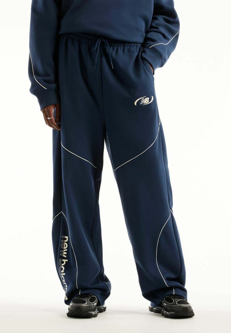 Personne portant un pantalon de survêtement New Balance bleu marine avec liseré blanc et baskets noires, debout avec une main dans la poche.