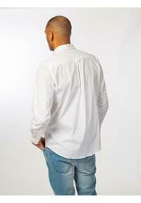 Camicia bianca con bottoni e maniche lunghe, caratterizzata da un dettaglio a piega sul retro, abbinata a jeans azzurri chiari. Tessuto morbido e design del colletto semplice.