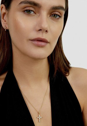 Woman with straight brown hair wears a black halter top, gold cross necklace with clear stones, and matching small cross earring.