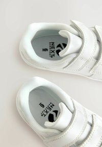 Chaussures blanches pour enfants avec une tige en cuir lisse, deux bandes velcro, un bout arrondi, et une semelle intérieure rembourrée affichant le logo de la marque.