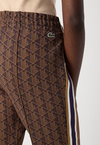 Pantaloni jogger marroni a disegno geometrico, con vita elastica e strisce laterali viola. Presentano un piccolo logo verde sulla coscia.