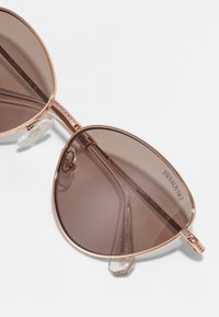 Óculos de sol com lentes grandes, de tonalidade castanha, armação de metal dourado e hastes com cristais. Inclui almofadas de nariz transparentes para maior conforto.