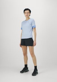 Lys blå kortermet skjorte med mørkere blå detaljer, kombinert med svarte shorts og svarte sportssko, satt mot en ensfarget bakgrunn.