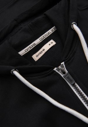 Felpa nera in cotone con zip e lacci bianchi, caratterizzata da un'etichetta con la scritta "ombre cotton line" e taglia M. Tessuto dalla consistenza liscia.