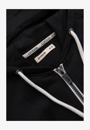 Sudadera de algodón negra con cremallera y cordones blancos, que presenta una etiqueta que dice "línea de algodón ombré" y tamaño M. Textura de tela lisa.